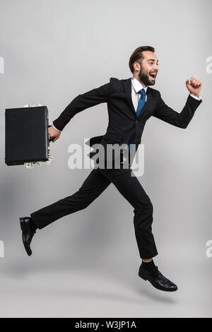 Full length of an attractive smiling young businessman wearing suit isolated over gray background, carrying briefcase, running Stock Photo