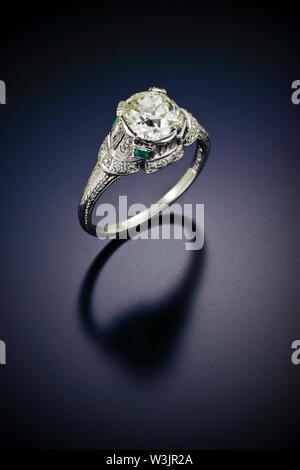 This antique diamond engagement ring with a one plus carat center stone and surrounded by accent side diamonds and emearld is set in engraved platinum Stock Photo