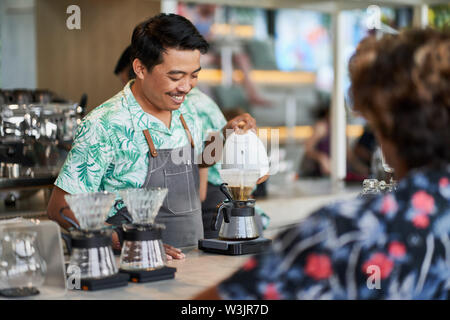 Candid lifestyle shot of smiling ethnic indonesian barista and small business owner preparing organic fair-trade coffee in bright trendy coffee shop Stock Photo