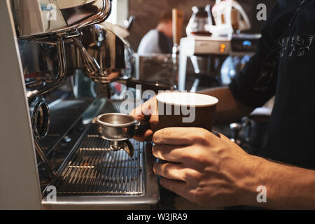 barista man holding hot coffee drink in paper cup in one hand and coffee holder in another standing near professional coffee machine in close up Stock Photo