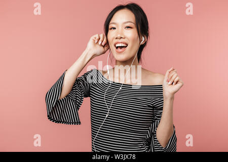 Photo of beautiful asian woman dressed in basic wear singing and listening to music with earphones isolated over red background in studio Stock Photo