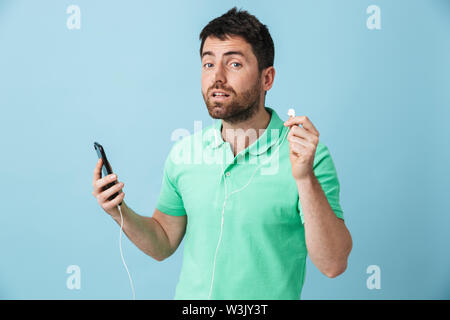 Image of a concentrated young handsome bearded man posing isolated over blue wall background using mobile phone listening music with earphones try to Stock Photo