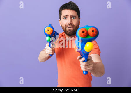 Image of a confused young handsome bearded man posing isolated over violet purple wall background holding water gun summer beach toy. Stock Photo