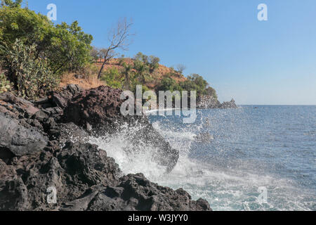 The sea surf breaks against a cliff of black lava boulders and water sprays high. Water drops fly through the air. The power of the sea. Stock Photo