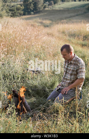 Man sitting in a grass beside a campfire on a meadow. Candid people, real moments, authentic situations Stock Photo
