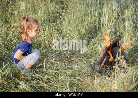 Little girl looking at campfire sitting in a grass. Candid people, real moments, authentic situations Stock Photo