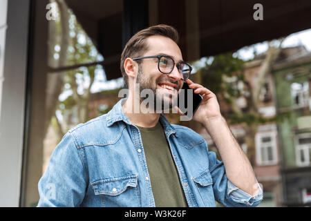 Photo of pleased young man wearing eyeglasses smiling and talking on smartphone while walking through city street Stock Photo