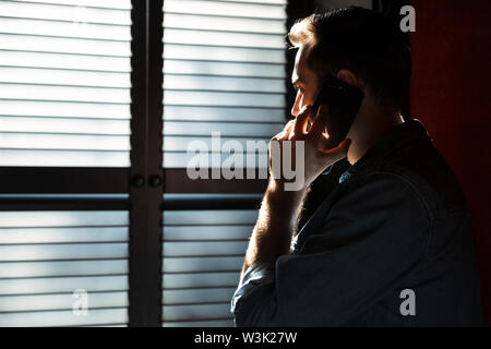 Silhouette of young attractive man talking on mobile phone while standing in dark room against the sunlight from window at shadow blinds Stock Photo