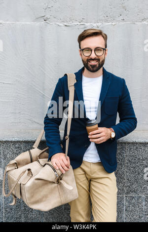 Portrait of bearded businessman wearing eyeglasses holding paper cup and carrying bag while standing near wall Stock Photo