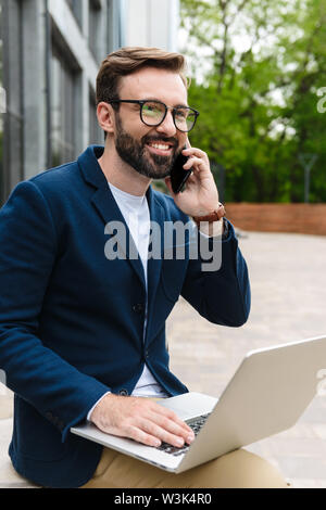 Portrait of bearded businessman wearing eyeglasses talking on cellphone and using laptop while sitting outdoors near building Stock Photo