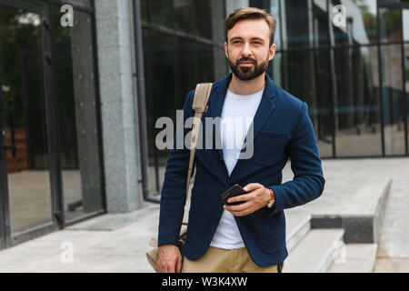 Attractive young bearded man wearing jacket walking outdoors at the street, carrying bag, holding mobile phone Stock Photo