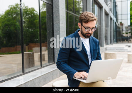 Attractive confident young bearded man wearing jacket working on laptop while sitting outdoors at the city Stock Photo