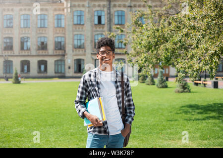 Smiling African American student with glasses and with books near college. Portrait of a happy black young man standing on a university background. Stock Photo
