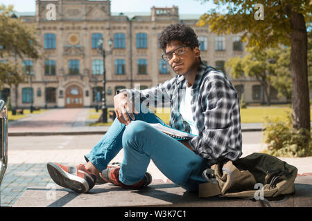 Smiling African American student with glasses and with books near college. Portrait of a happy black young man standing on a university background. Stock Photo