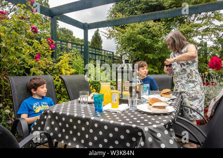 A family eat hamburgers sat at a table and chairs under a pergola outside on a garden Stock Photo