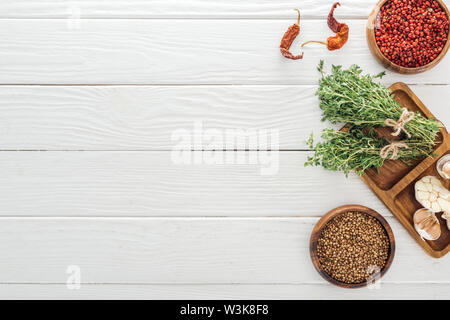 top view of dried chili peppers, coriander and pink peppercorn in bowls near board with garlic cloves and thyme on white wooden table Stock Photo