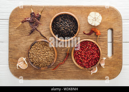 top view of black pepper, coriander and pink peppercorn in bowls on wooden chopping board Stock Photo