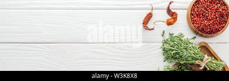 panoramic shot of dried chili peppers, pink peppercorn in bowl near board with thyme on white wooden table Stock Photo