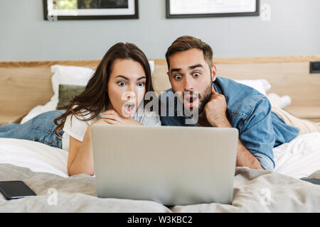Beautiful shocked young couple in love at home, laying in bed, looking at laptop computer Stock Photo