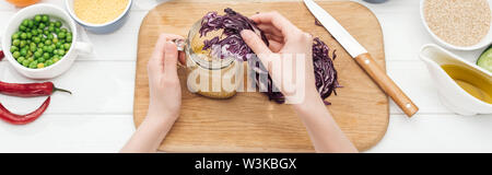 cropped view of woman adding cut red cabbage in jar with salad on wooden white table, panoramic shot Stock Photo