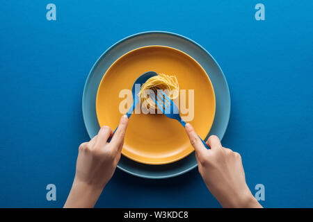 cropped view of woman holding blue spoon and taking vermicelli pasta with fork on yellow plate above another plate on blue background Stock Photo