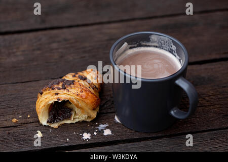 Hot chocolate in a blue-grey ceramic mug next to a half of chocolate croissant isolated on rustic dark brown wood table. Stock Photo
