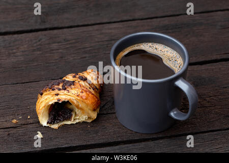Black coffee in a blue-grey ceramic mug next to a half of chocolate croissant isolated on rustic dark brown wood table. Stock Photo