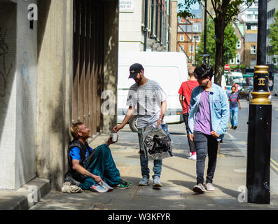 Two young guys giving some money to a homeless person which is sitting on the ground in the street Stock Photo