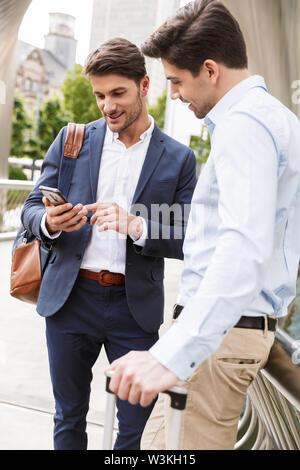 Photo of a pleased happy young two friends colleagues business men standing outdoors talking with each other using mobile phone. Stock Photo