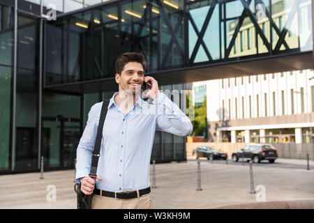 Image of a smiling pleased young businessman walking by street near business center talking by mobile phone holding backpack. Stock Photo