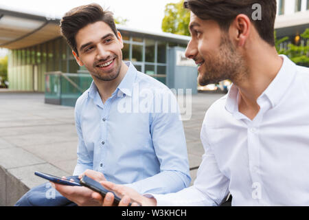 Image of a cheerful positive young two friends colleagues business men sitting near business center talking with each other using mobile phones. Stock Photo