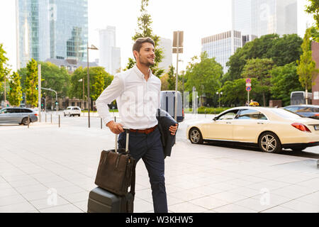 Image of a young pleased smiling happy businessman walking outdoors on the street with suitcase holding jacket. Stock Photo