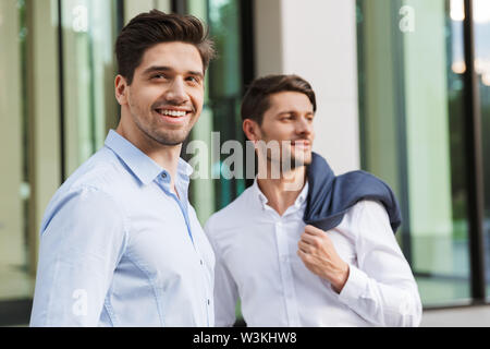 Photo of a young smiling pleased two colleagues businessmen walking outdoors on the street. Stock Photo