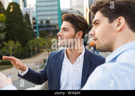 Photo of a handsome serious unshaved two friends colleagues business men outdoors on the street talking with each other looking aside. Stock Photo