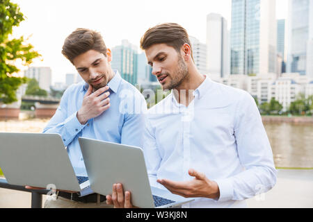 Photo of a young concentrated two colleagues businessmen standing outdoors on the street with beautiful sunlight over lake background using laptop com Stock Photo
