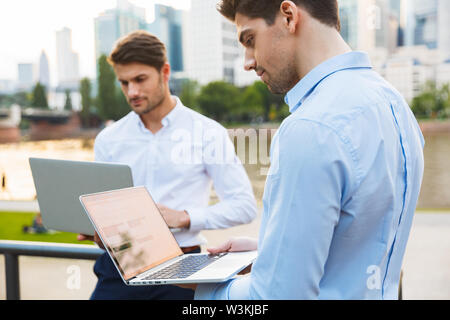Photo of a young two serious colleagues businessmen standing outdoors on the street with beautiful sunlight over lake background using laptop computer Stock Photo