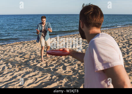 Two happy young men playing with frisbee at the beach, close up back view of a man Stock Photo