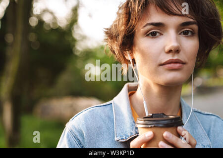 Image of thoughtful caucasian woman 20s wearing denim jacket drinking takeaway coffee and using earphones while walking through green park Stock Photo