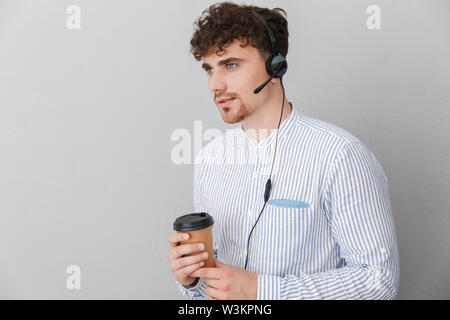 Portrait of young beautiful phone operator man wearing microphone headset and holding coffee cup while speaking with client isolated over gray backgro Stock Photo