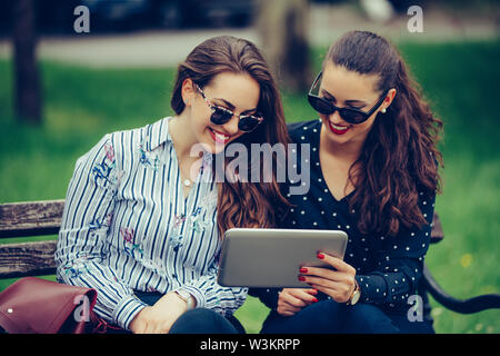 Two beautiful women laughing watching media content together in a digital tablet, sitting on a bench in the park.