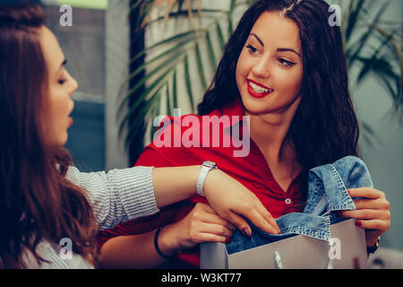 Two young women sharing their new purchases with each other. They having coffee break after shopping. Consumerism, shopping, lifestyle concept. Stock Photo