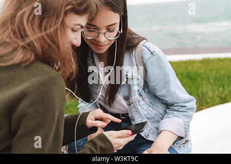 Cropped photo of a smiling emotional young pretty friends women students walking outdoors listening music with earphones using mobile phone. Stock Photo
