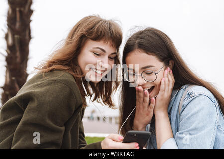 Image of a shocked surprised emotional young pretty friends women students walking outdoors listening music with earphones using mobile phone. Stock Photo