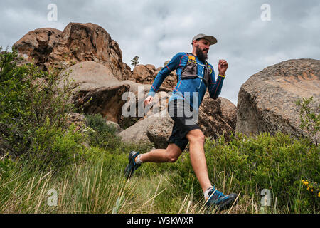 Male runner running on a mountain trail. Athlete runs in the mountains among the rocks. Man in blue jersey and black shorts training outdoors Stock Photo