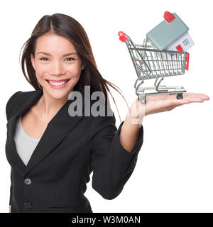 Real estate agent selling home holding mini house in shopping cart. Female realtor in business suit showing model house smiling happy isolated on white background. Multiracial Asian woman agent. Stock Photo