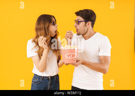 Portrait of surprised couple man and woman in 3D glasses rejoicing while standing together with popcorn bucket isolated over yellow background Stock Photo