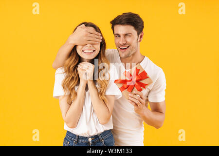 Portrait of happy couple in basic t-shirts standing together while man holding present box and covering woman's eyes isolated over yellow background Stock Photo