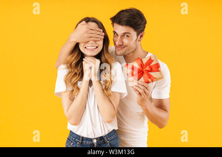 Portrait of caucasian couple in basic t-shirts standing together while man holding present box and covering woman's eyes isolated over yellow backgrou Stock Photo