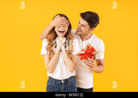 Portrait of attractive couple in basic t-shirts standing together while man holding present box and covering woman's eyes isolated over yellow backgro Stock Photo