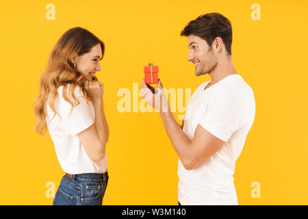 Portrait of beautiful couple standing together while handsome man making proposal to happy woman with ring box isolated over yellow background Stock Photo